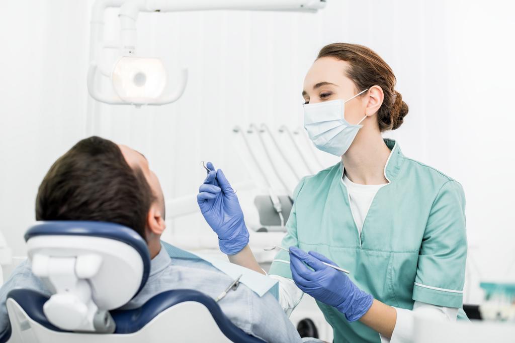 Naispuolinen hammaslääkäri maskissa pitäen hammasinstrumentteja lähellä potilasta hammashoitolassa
 - Valokuva, kuva