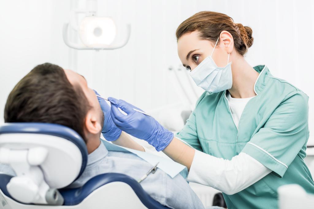 vrouwelijk tandarts in masker tandheelkundige instrumenten te houden terwijl het werken met de patiënt in tandheelkundige kliniek - Foto, afbeelding