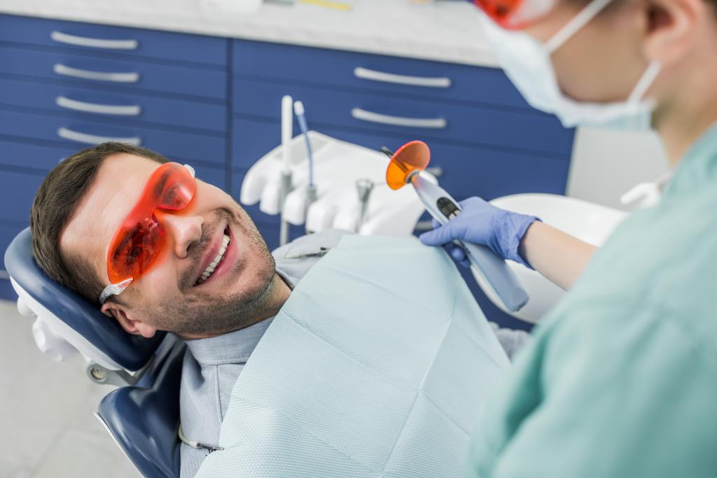 messa a fuoco selettiva di uomo allegro in occhiali vicino dentista tenendo strumento dentale prima della procedura di sbiancamento
  - Foto, immagini