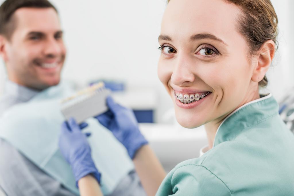 selective focus of cheerful dentist with braces on teeth smiling while holding color palette near patient  - Photo, Image