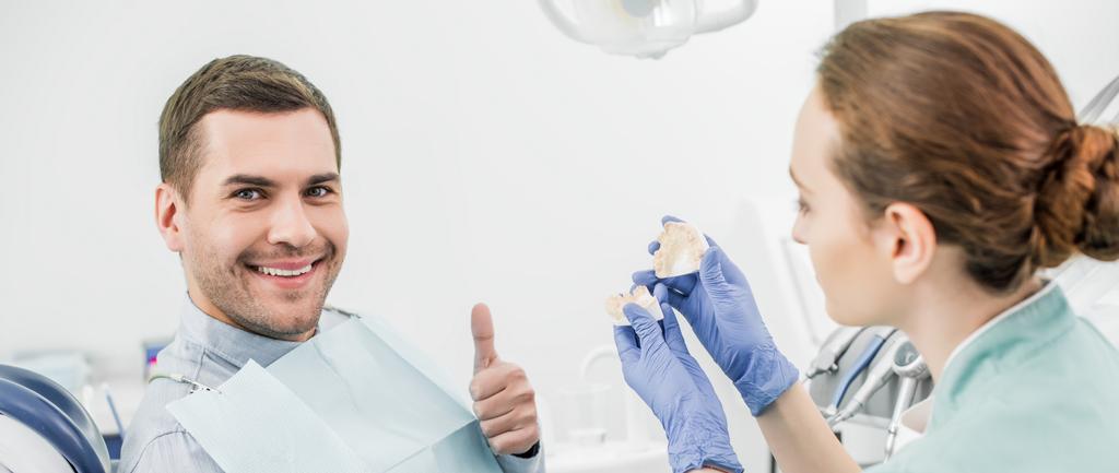 selective focus of handsome man showing thumb up near female dentist holding teeth model  - Photo, Image