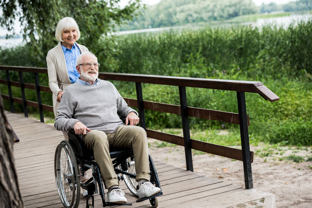 šťastný starší muž na invalidním vozíku se usmívající se ženou na dřevěný most v parku - Fotografie, Obrázek