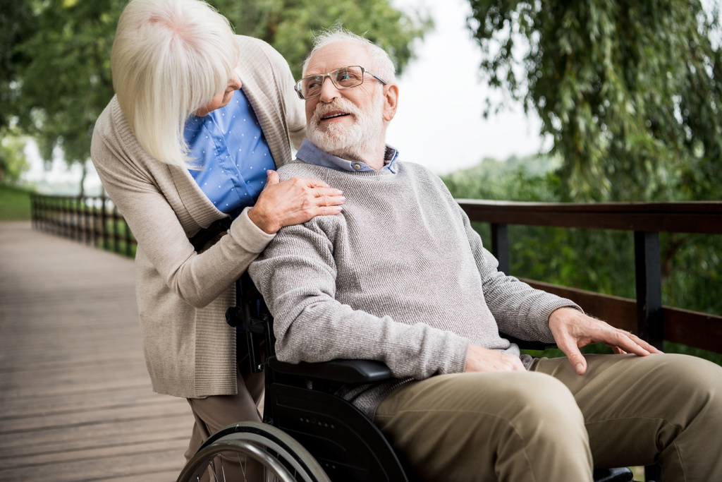 senior woman looking at smiling husband in wheelchair while holding hand on his shoulder - Photo, Image