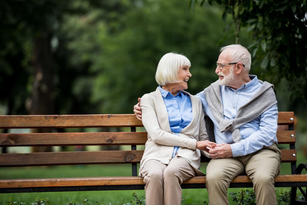 happy senior couple holding hands and looking at each other while sitting on wooden bench park - Photo, Image