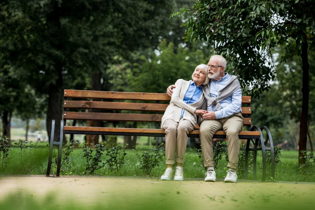 シニア カップルを抱きしめると公園の木製ベンチで休んでいる間手を繋いでいる笑顔 - 写真・画像