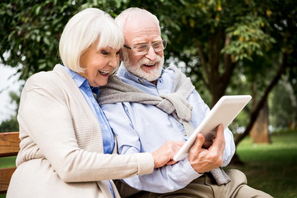 sonriente pareja de ancianos utilizando tableta digital mientras está sentado en el banco en el parque
 - Foto, imagen