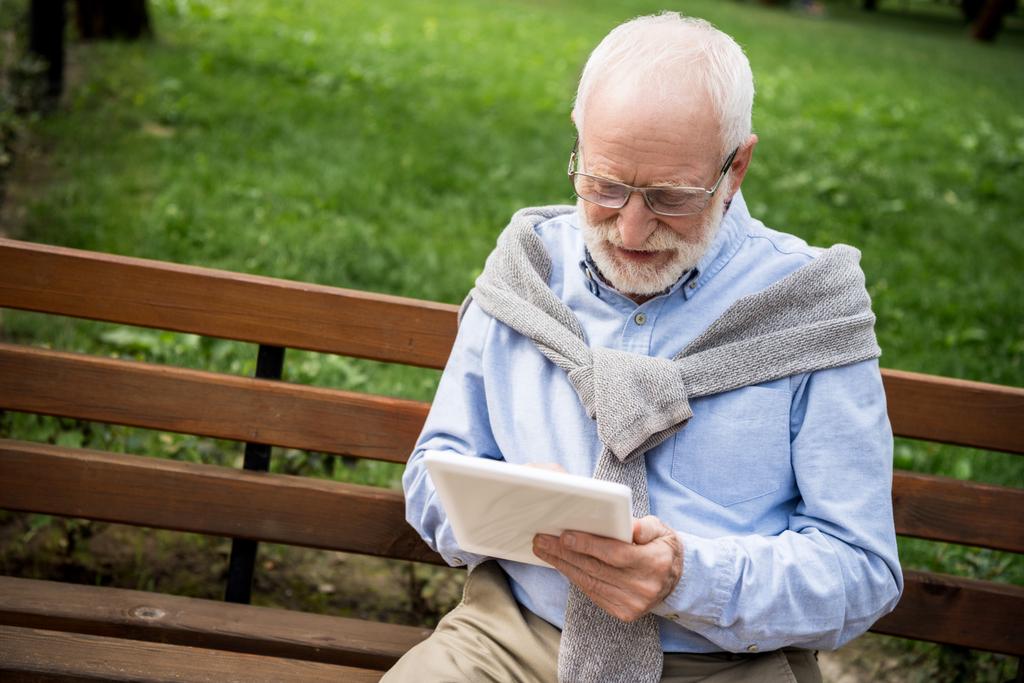 glimlachend senior man met digitale tablet terwijl zittend op houten bankje - Foto, afbeelding