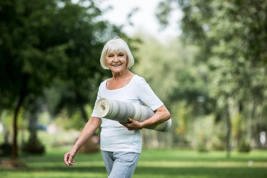 счастливая пожилая женщина с фитнес-ковриком, гуляющая в парке
 - Фото, изображение