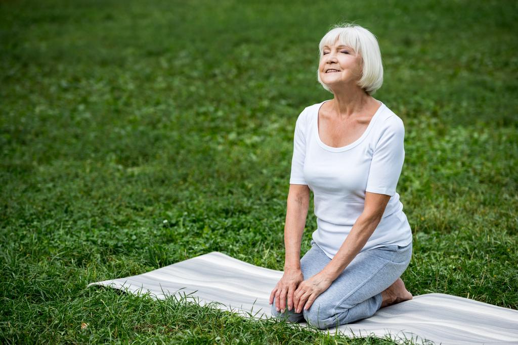 szczęśliwy senior kobieta siedzi w medytacji pozie z ręce na kolanach i zamknięte oczy - Zdjęcie, obraz