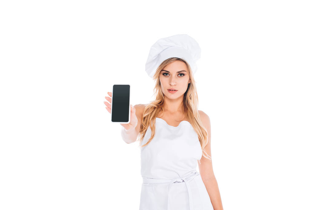 aantrekkelijke blonde cook vrouw in uniform bedrijf smartphone met leeg scherm geïsoleerd op wit - Foto, afbeelding