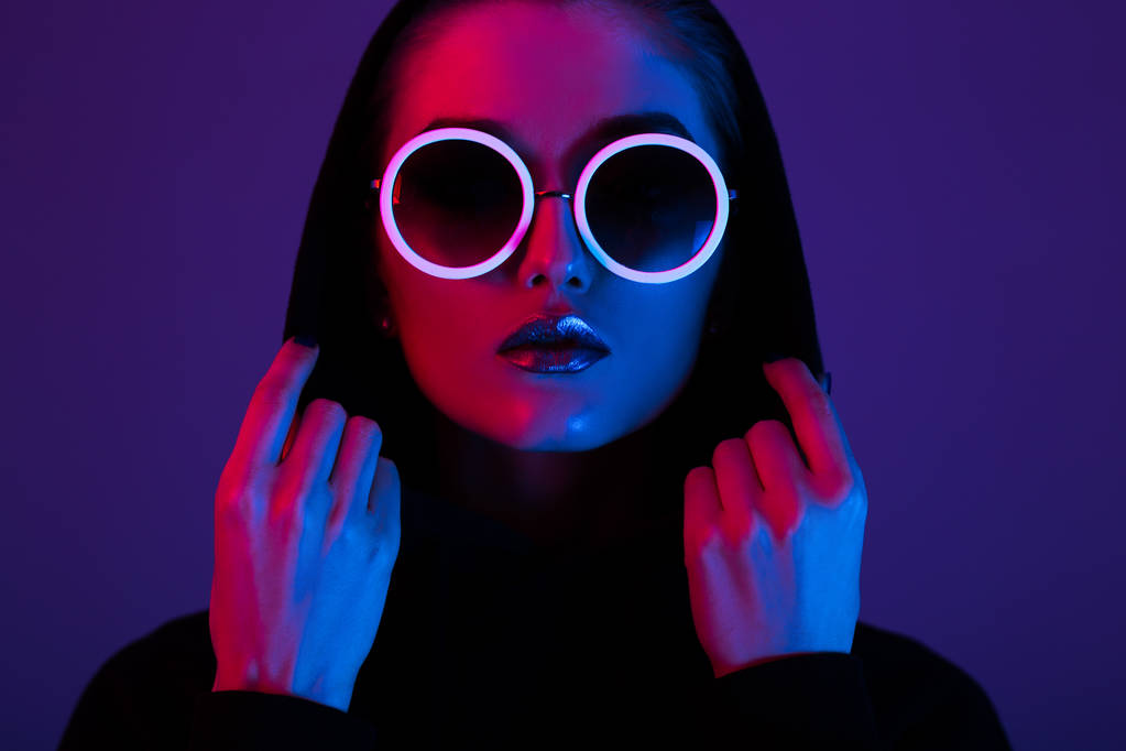 Siyah kazaklı bir kukuleta ile ve güneş gözlüğü kırmızı ve mavi neon ışık stüdyo yuvarlak genç kız moda portresi - Fotoğraf, Görsel