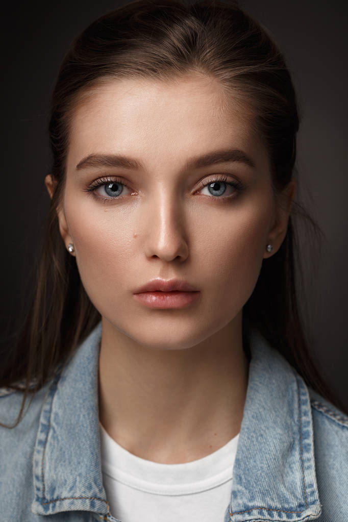 Portrait of beautiful brunette girl with hair tied back dressed in jeans jacket on the dark background in the studio - Photo, Image