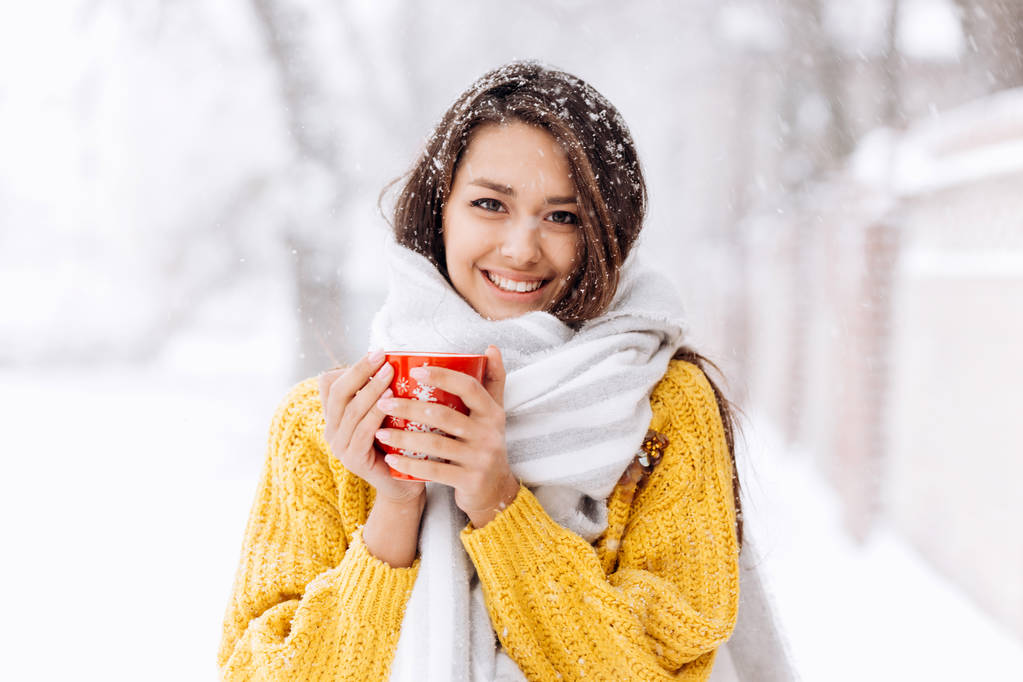 Usmívající se černovlasá dívka v žlutém svetru, džíny a bílou šátek stojí s červený hrnek na zasněžené ulici na zimní den - Fotografie, Obrázek