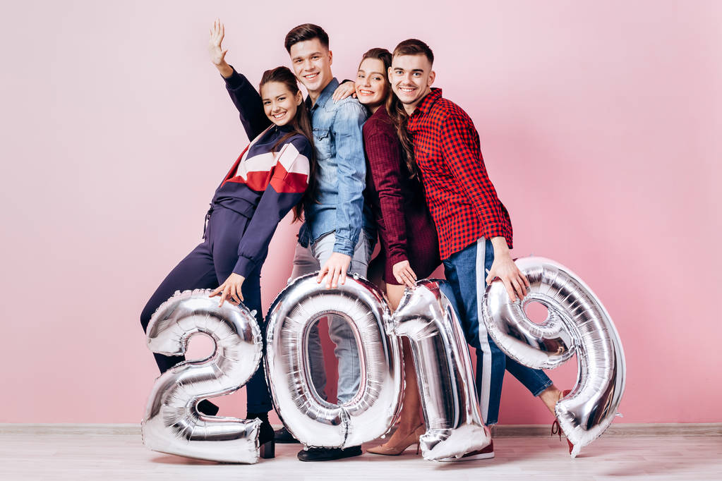Feliz companhia de duas meninas e dois caras vestidos com roupas elegantes estão segurando balões na forma de números 2019 em um fundo rosa no estúdio
 - Foto, Imagem