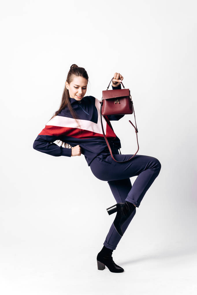 Girl dressed in sporty blue suit with a red and white print on a sweatshirt and heels poses with bag in her hands on the white background in the studio - Photo, Image