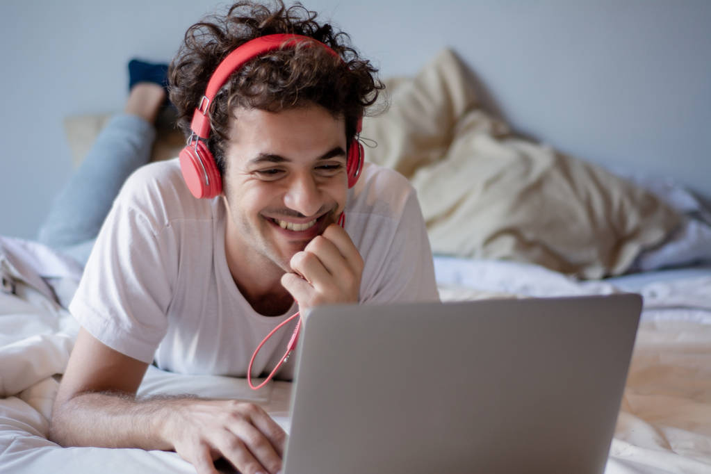 Feliz joven escuchando música con auriculares y usando un portátil en su cama. En interiores
 - Foto, Imagen