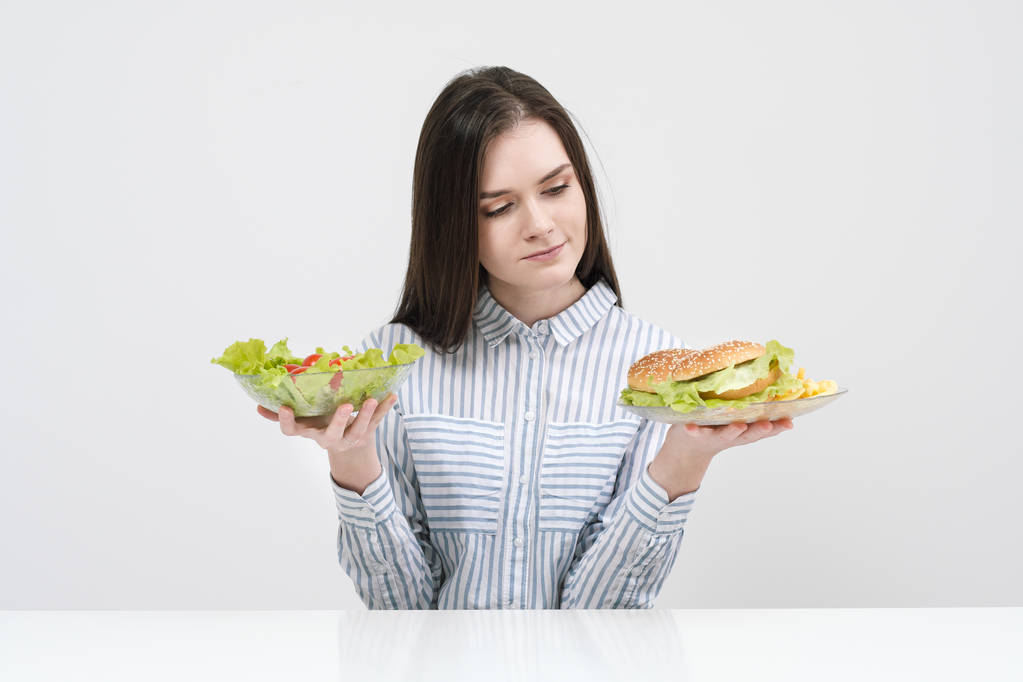 Szczupła brunetka dziewczyna na białym tle wybiera między płyty fast food i hamburgery i zdrowe jedzenie Sałatka. - Zdjęcie, obraz