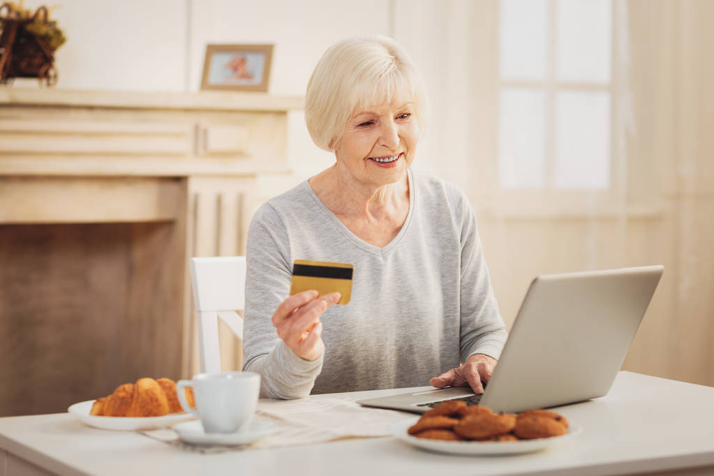 Online αγορές. Σύγχρονη χαμογελώντας συνταξιούχος αίσθημα απασχολημένος ψωνίζοντας online έχοντας νόστιμο γαλλικό πρωινό - Φωτογραφία, εικόνα
