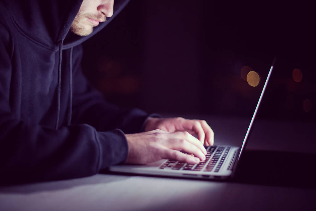 Молодой талантливый хакер использует ноутбук, работая в темном офисе с большим городским освещением на заднем плане ночью - Фото, изображение