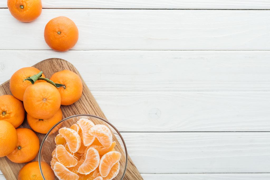 vista superior de fatias de tangerina descascadas em tigela de vidro e tangerinas maduras inteiras em tábua de corte de madeira
  - Foto, Imagem