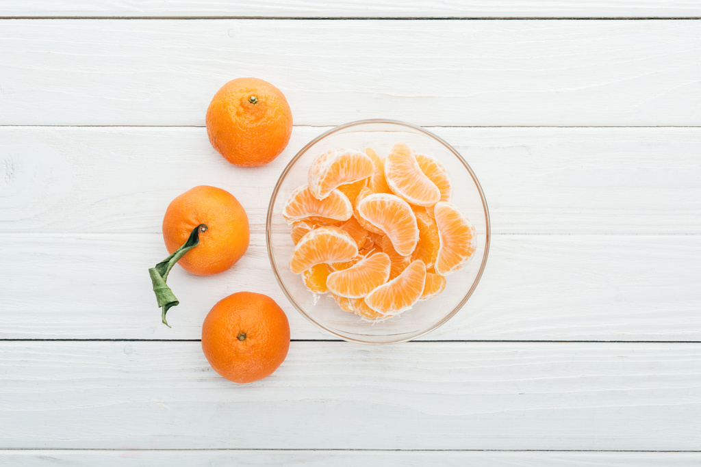pohled shora oloupaná mandarinka výsečí skleněná mísa a zcela zralé mandarinky kolem na dřevěné bílé pozadí - Fotografie, Obrázek