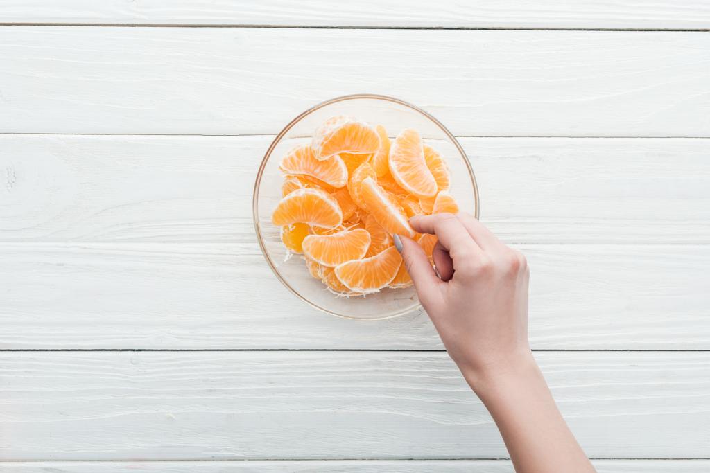 vista recortada de la mujer tomando rebanada de mandarina de tazón de vidrio sobre fondo blanco de madera
 - Foto, Imagen