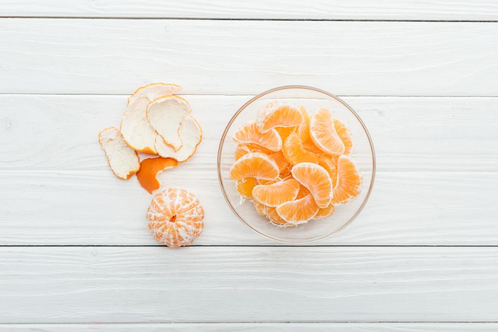 vista superior de fatias de tangerina descascadas em tigela de vidro e casca no fundo branco de madeira
 - Foto, Imagem