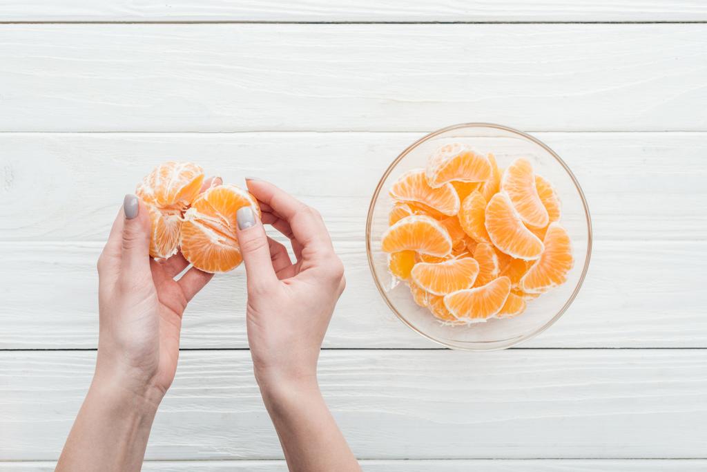 cropped view of woman holding peeled tangerine near glass bowl with tangerine slices on wooden white background - Photo, Image