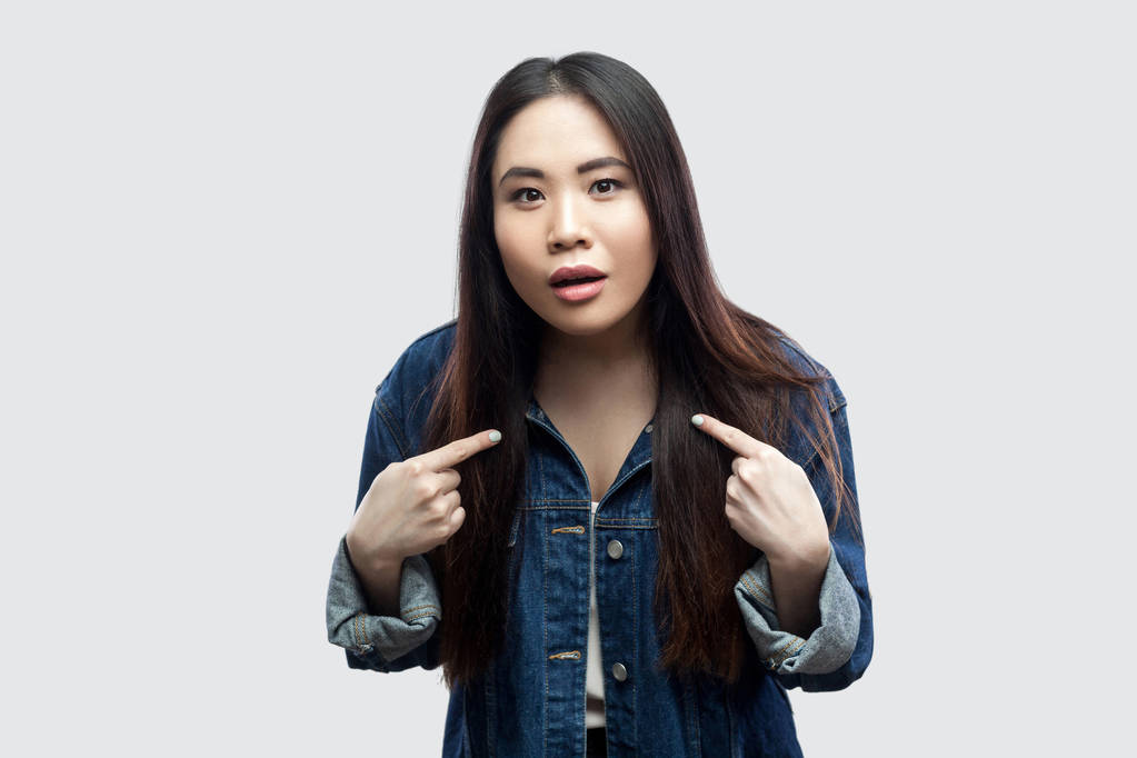 shocked beautiful brunette asian young woman in casual blue denim jacket pointing herself and looking at camera on light grey background. - Photo, Image