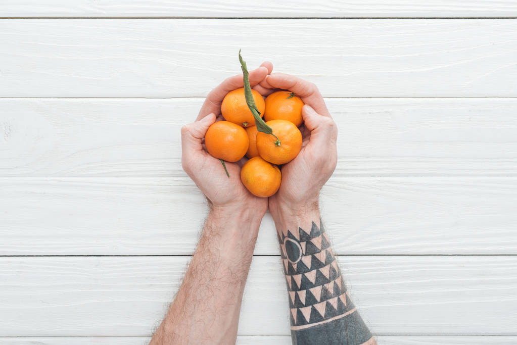 weergave van de getatoeëerde mens houden hoop mandarijnen op witte houten oppervlak bijgesneden - Foto, afbeelding