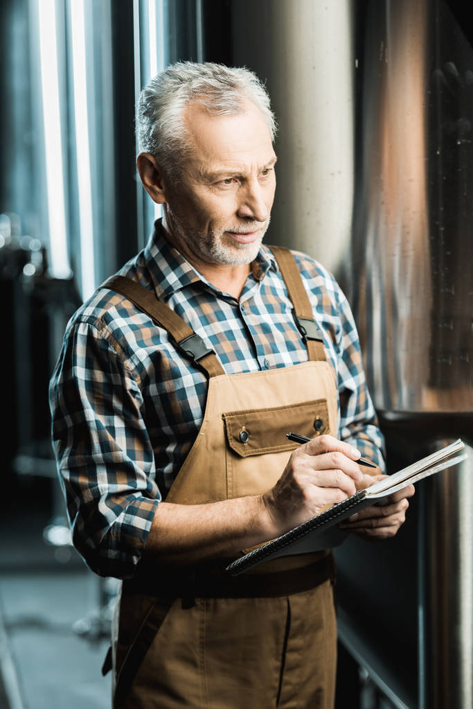 профессиональный пивовар-мужчина в рабочем комбинезоне пишет в блокноте на пивоварне
 - Фото, изображение