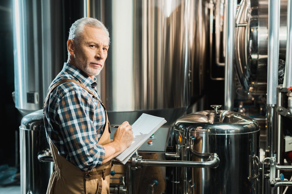 Senior männlicher Brauer schreibt in Notizblock, während er Brauereiausrüstung überprüft - Foto, Bild