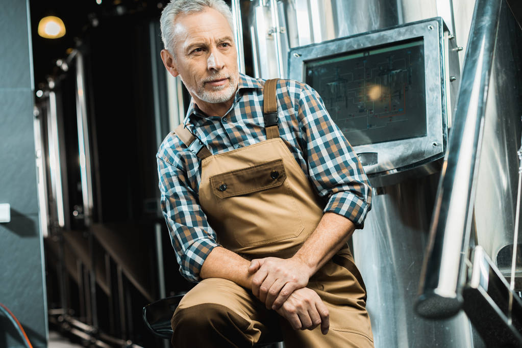 старший чоловічий пивовар в робочому комбінезоні, сидячи в пивоварні
 - Фото, зображення