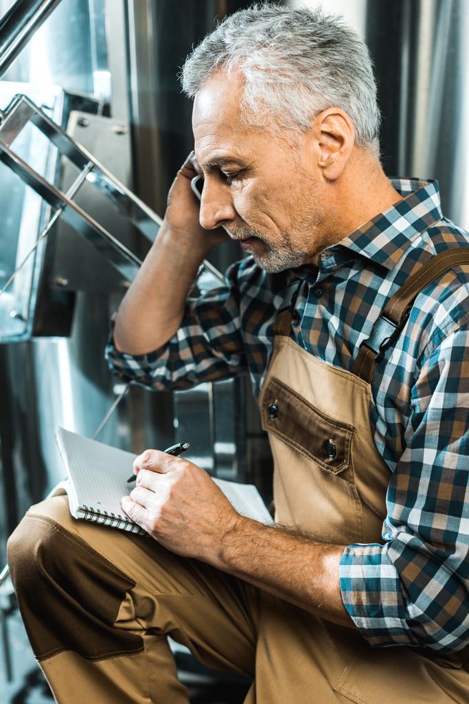 Braumeister in Arbeitshose spricht auf Smartphone, während er in Brauerei-Notizblock schreibt - Foto, Bild