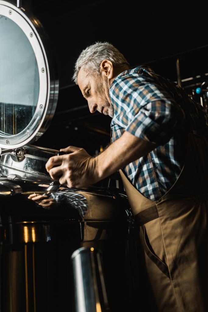 Senior mannelijke brouwer in werkende Amerikaanse overalls brouwerij apparatuur controleren - Foto, afbeelding