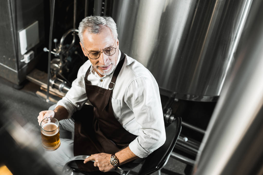vista aérea de cervecero senior en delantal sentado en silla y sosteniendo un vaso de cerveza en la cervecería
 - Foto, Imagen