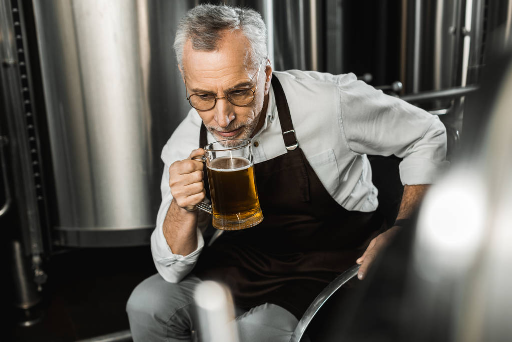 старший пивовар в фартусі сидить на стільці і тестує пиво в пивоварні
 - Фото, зображення