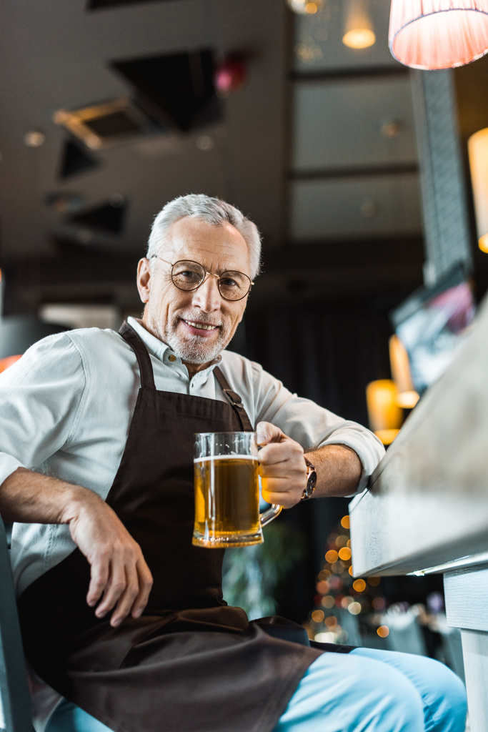 senior worker in apron holding glass of beer near bar counter - Photo, Image