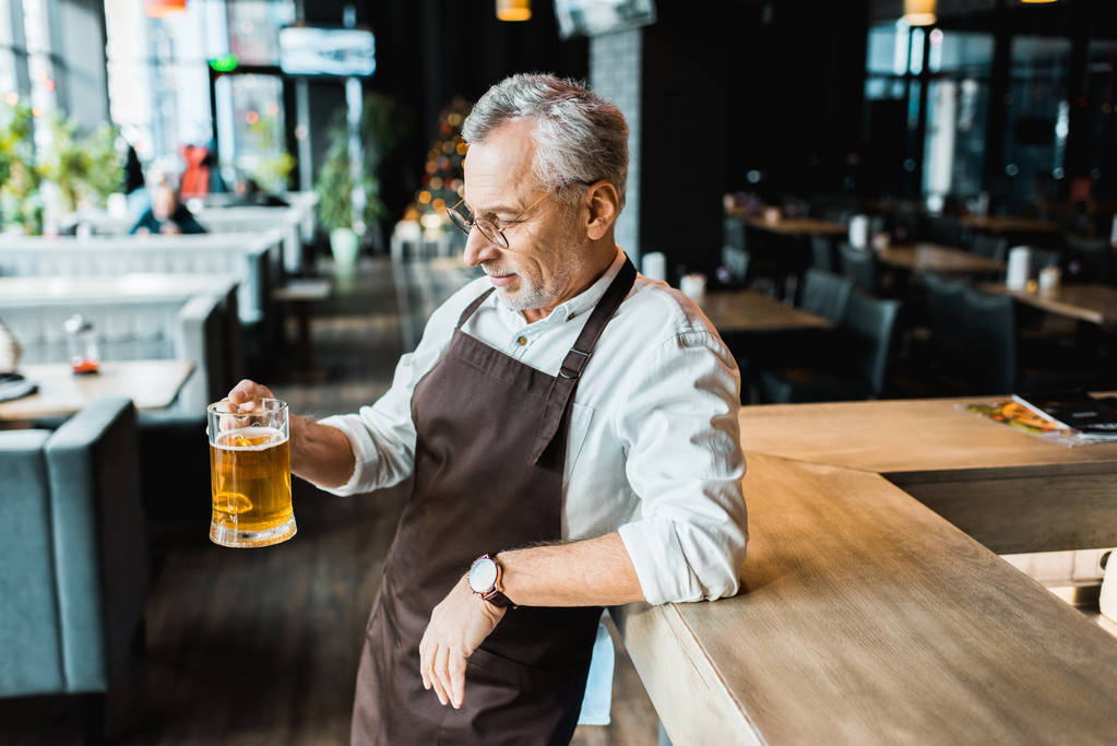 Senior werknemer in schort holding glas bier in de kroeg - Foto, afbeelding