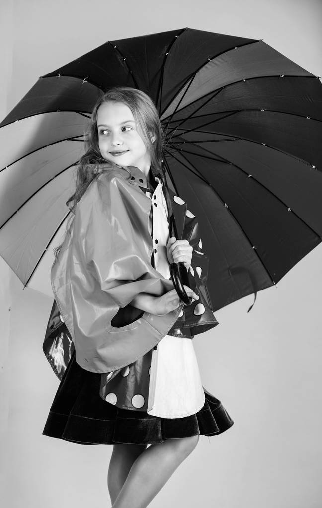 Ευτυχισμένο παιδί κορίτσι κρατήστε φθορά ομπρέλα αδιάβροχο μανδύα. Αδιάβροχο αξεσουάρ κάνουν βροχερή μέρα χαρούμενη και ευχάριστη. Εμπιστοσύνη σε αυτήν εμπίπτουν ενδύματα. Αδιάβροχο αξεσουάρ κατασκευής - Φωτογραφία, εικόνα