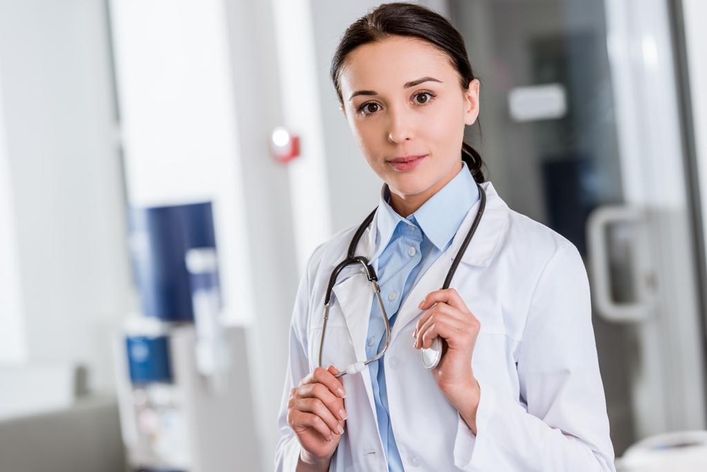 Young doctor in white coat touching stethoscope and looking at camera - Photo, Image