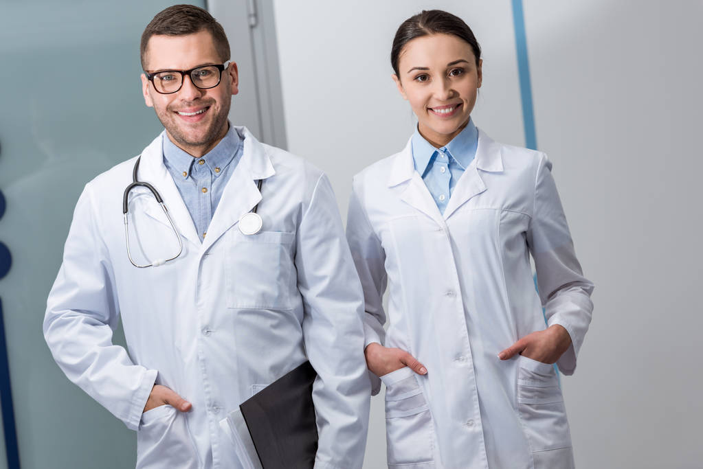Szczęśliwy lekarzy z folderu i stetoskop stojący z rękami w kieszeniach - Zdjęcie, obraz
