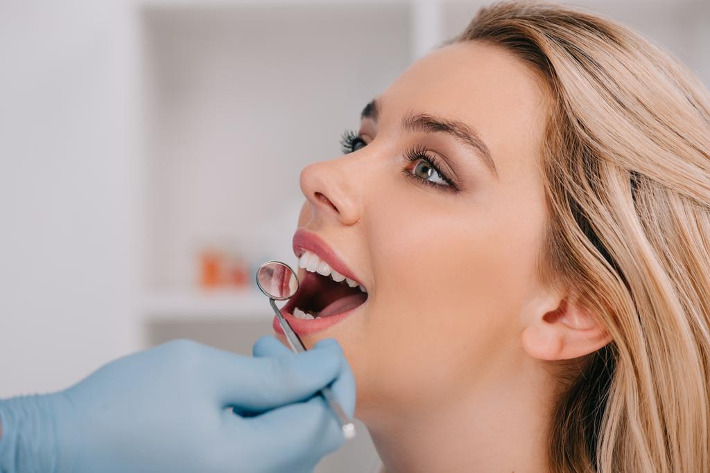 ο οδοντίατρος εξετάζει τα δόντια της νεαρής γυναίκας με στόμα καθρέφτη - Φωτογραφία, εικόνα