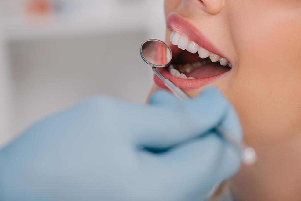 обрізаний вид стоматолога, який вивчає зуби молодої жінки з дзеркалом рота
 - Фото, зображення