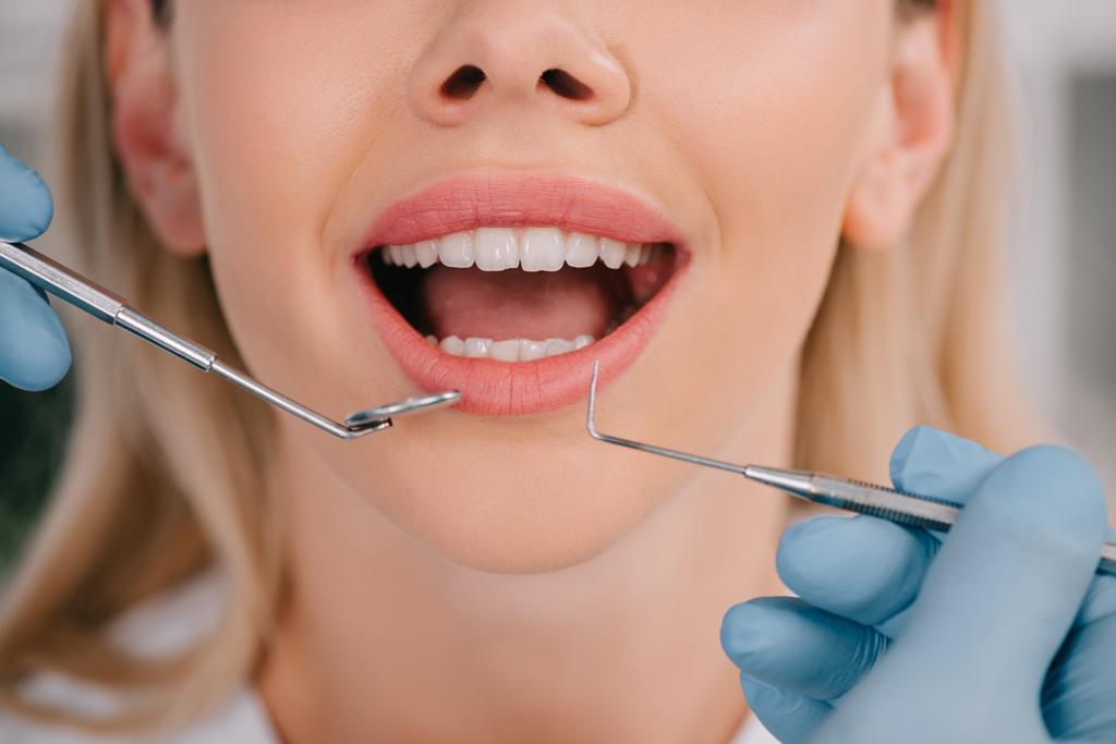 обрізаний вид на стоматолога, який вивчає зуби молодої жінки з дзеркалом рота та стоматологічним зондом
 - Фото, зображення