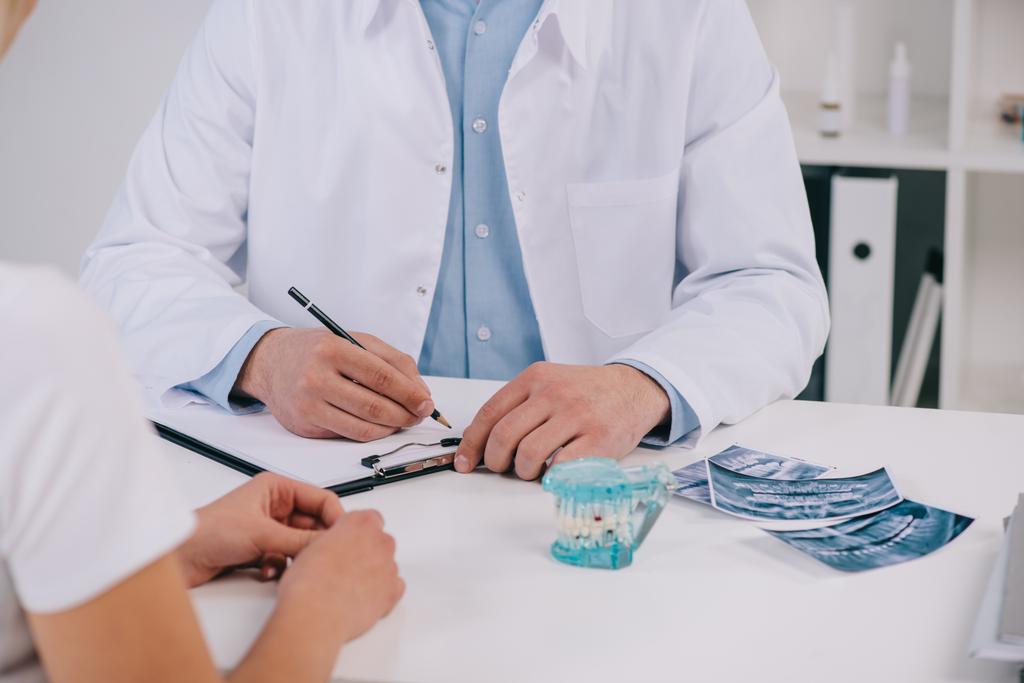 bijgesneden weergave van orthodontist diagnose schrijven tijdens overleg met vrouw in kliniek - Foto, afbeelding