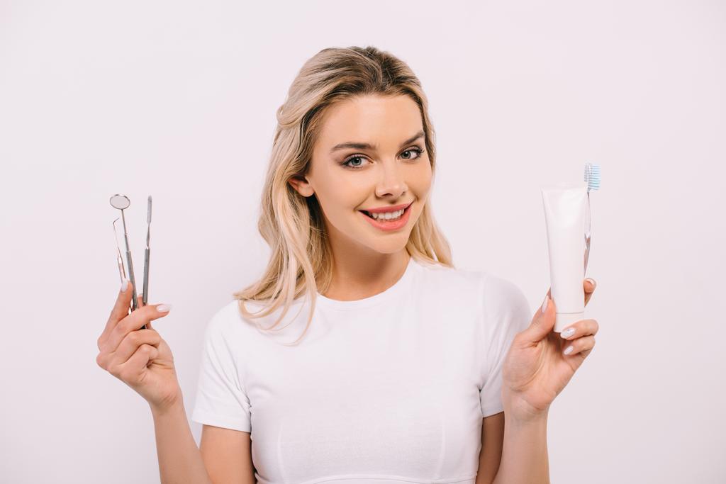 bela mulher sorridente olhando para a câmera enquanto segurando pasta de dentes, escova de dentes e instrumentos dentários isolados em branco
 - Foto, Imagem