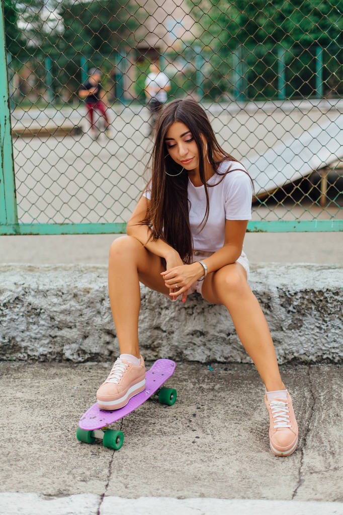 Nyári életmód kép trendi csinos fiatal lány ül a gördeszka coart vele lila műanyag gördeszka. - Fotó, kép