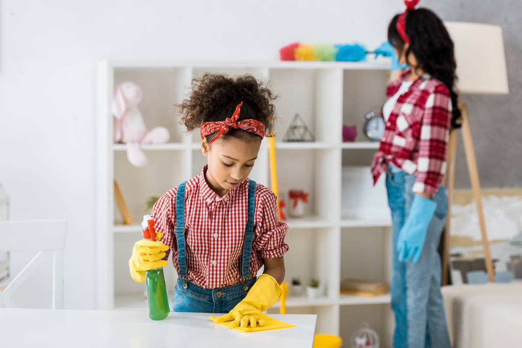 selectieve aandacht van schattige Afro-Amerikaanse kind tabel terwijl moeder afstoffen meubilair schoonmaken - Foto, afbeelding