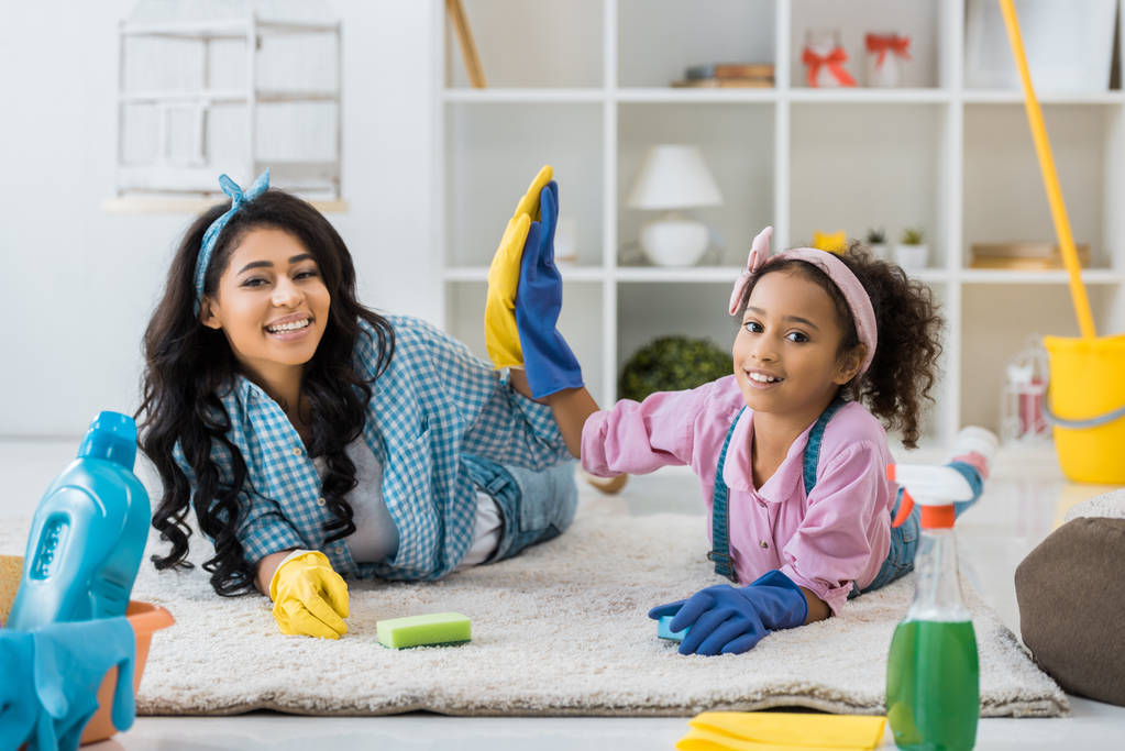 Smling african american mother and child in bright rubber gloves giving high five while lying on carpet
 - Фото, изображение